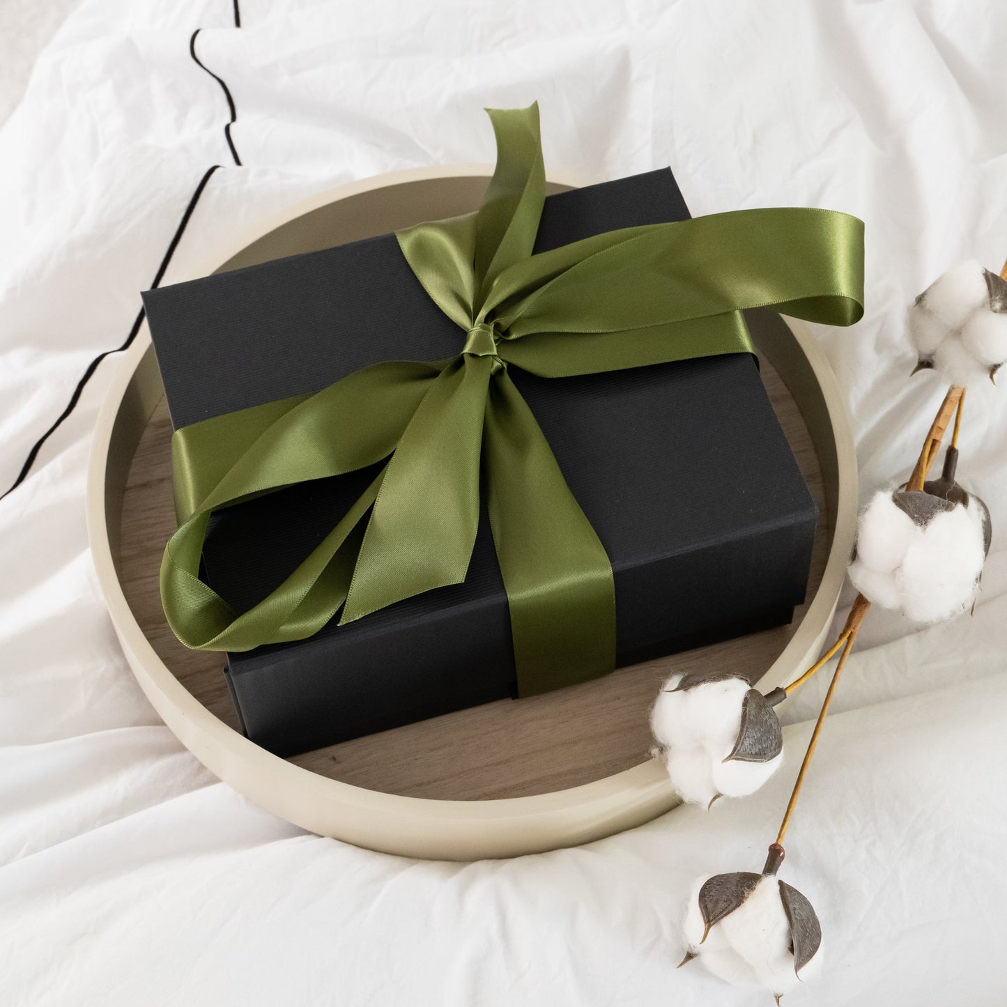 Black gift box with a green ribbon on a white surface with cotton flowers.