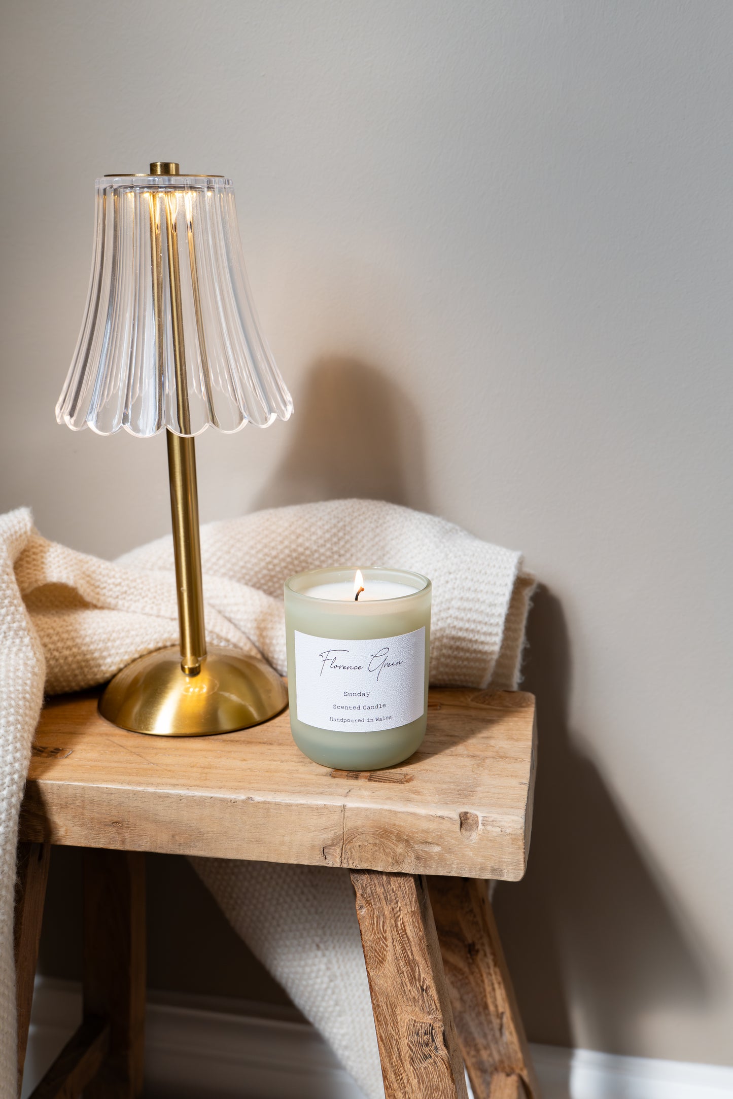 Candle with a label on a wooden stool next to a gold lamp against a neutral wall.