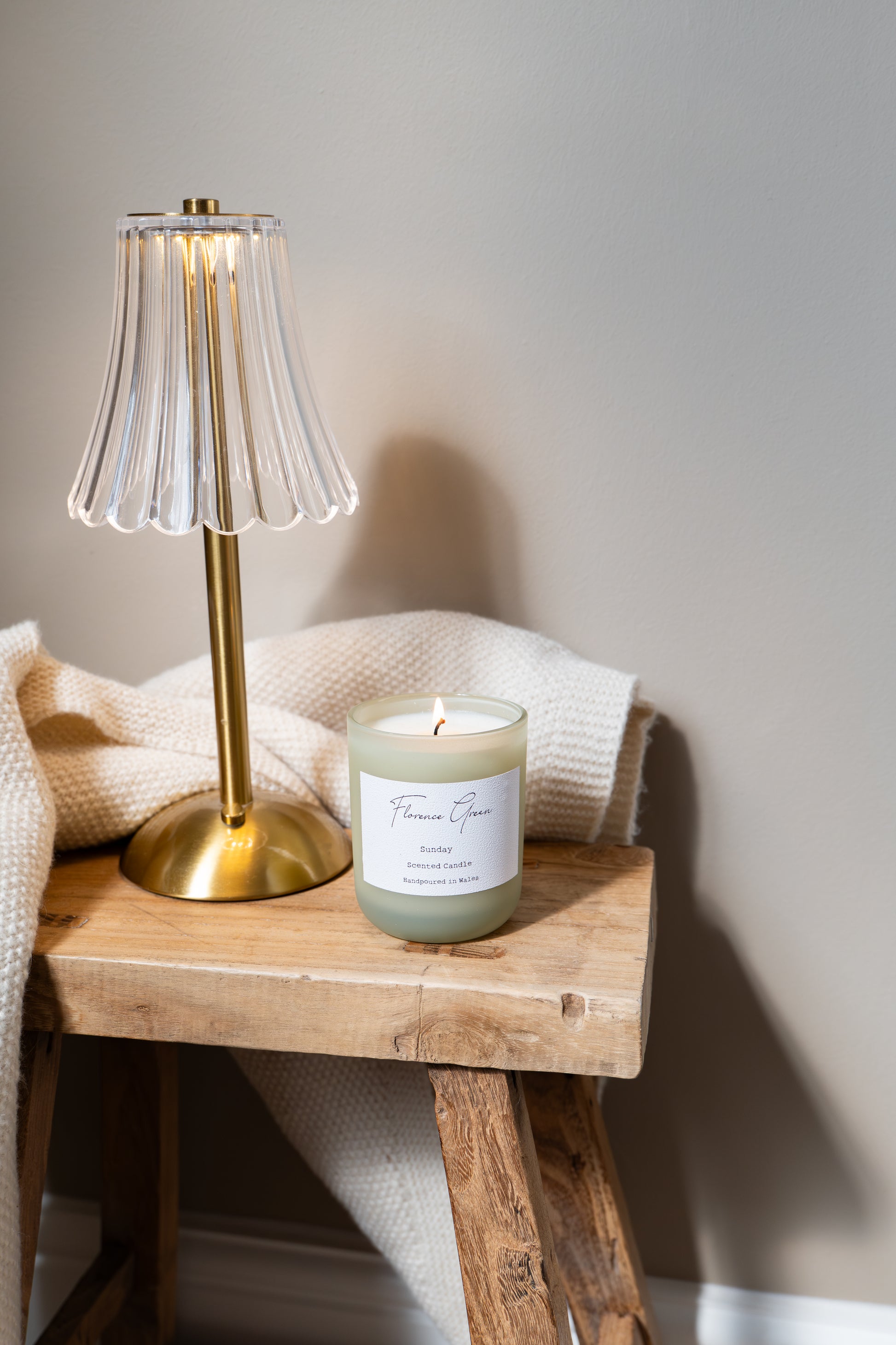 Candle with a label on a wooden stool next to a gold lamp against a neutral wall.
