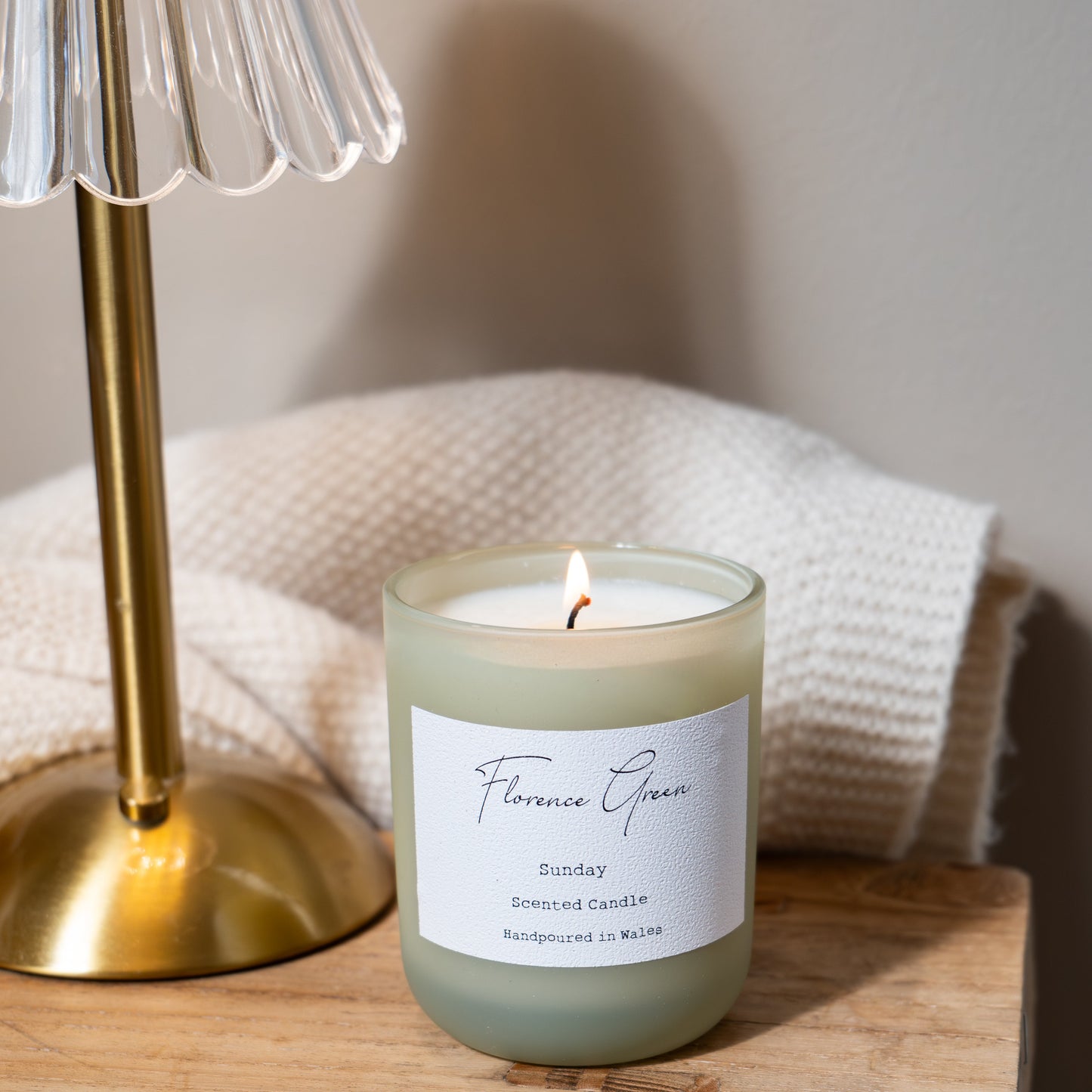 Candle with a label on a wooden surface next to a gold lamp.