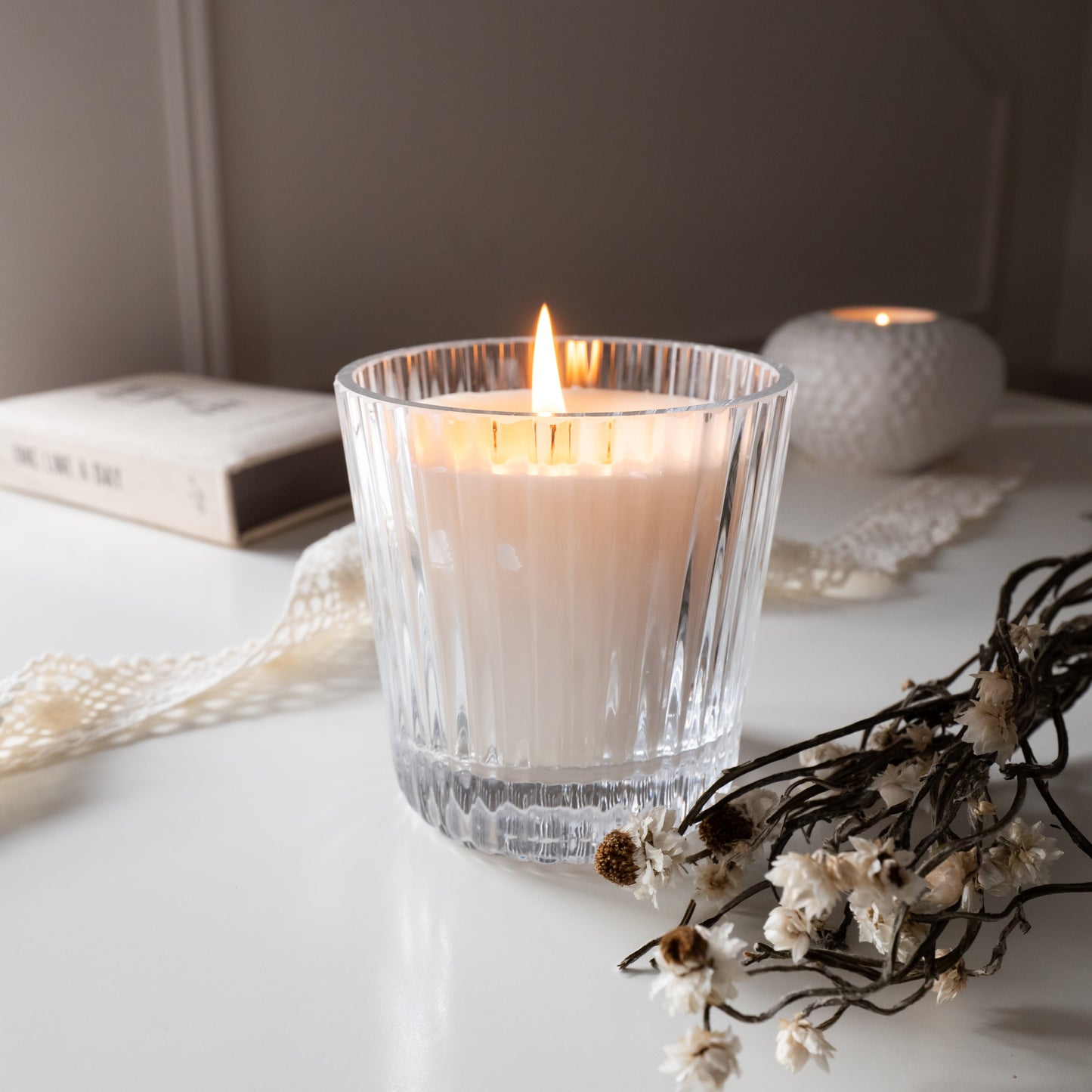 Candle in a glass holder on a white surface with flowers and a book in the background